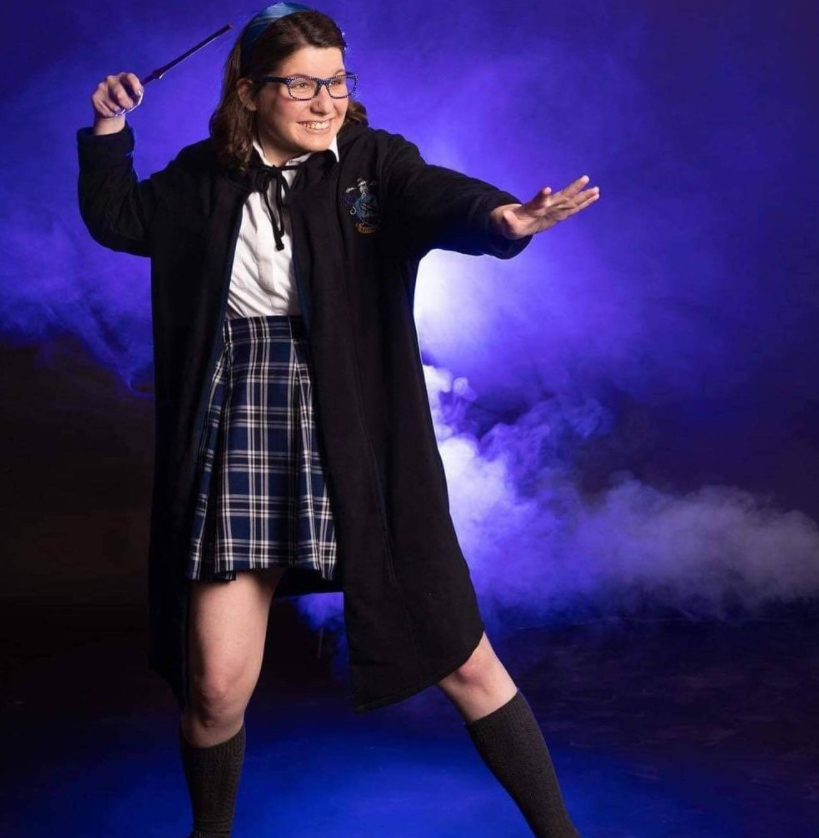 Amy is wearing a short black cape, white shirt, and blue plaid skirt. She is holding a wand above her head. The background is deep blue with white mist.
