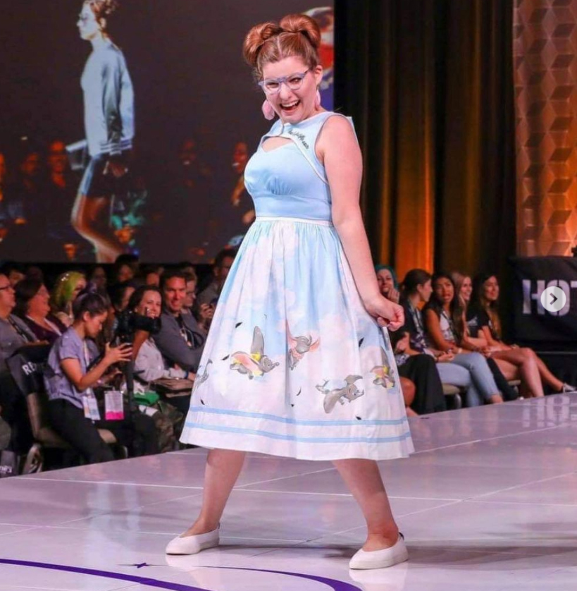 Amy is walking on a white fashion show runway. She has her hair in two buns, is wearing light blue glasses and pink feather earrings. Her dress is bule with images of Dumbo the elephant at the bottom.