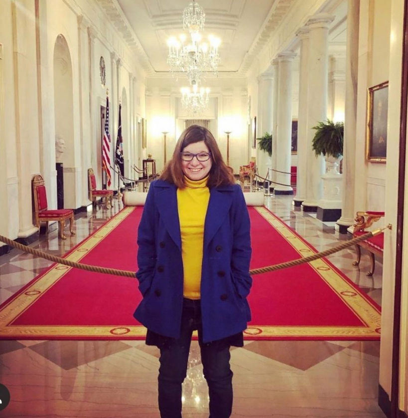 mage of Amy in White House Cooridor. She is a Caucasian young woman, wearing glasses, with shoulder length brown hair. She is wearing a yellow turtleneck top with bright blue jacket. Behind her is a long red carpet. Above her are chandeliers.